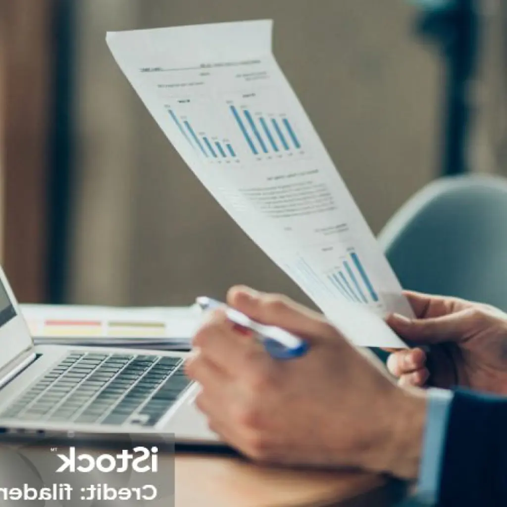 Nahaufnahme eines Laptops, davor eine Hand, die ein Blatt mit Graphen enthält