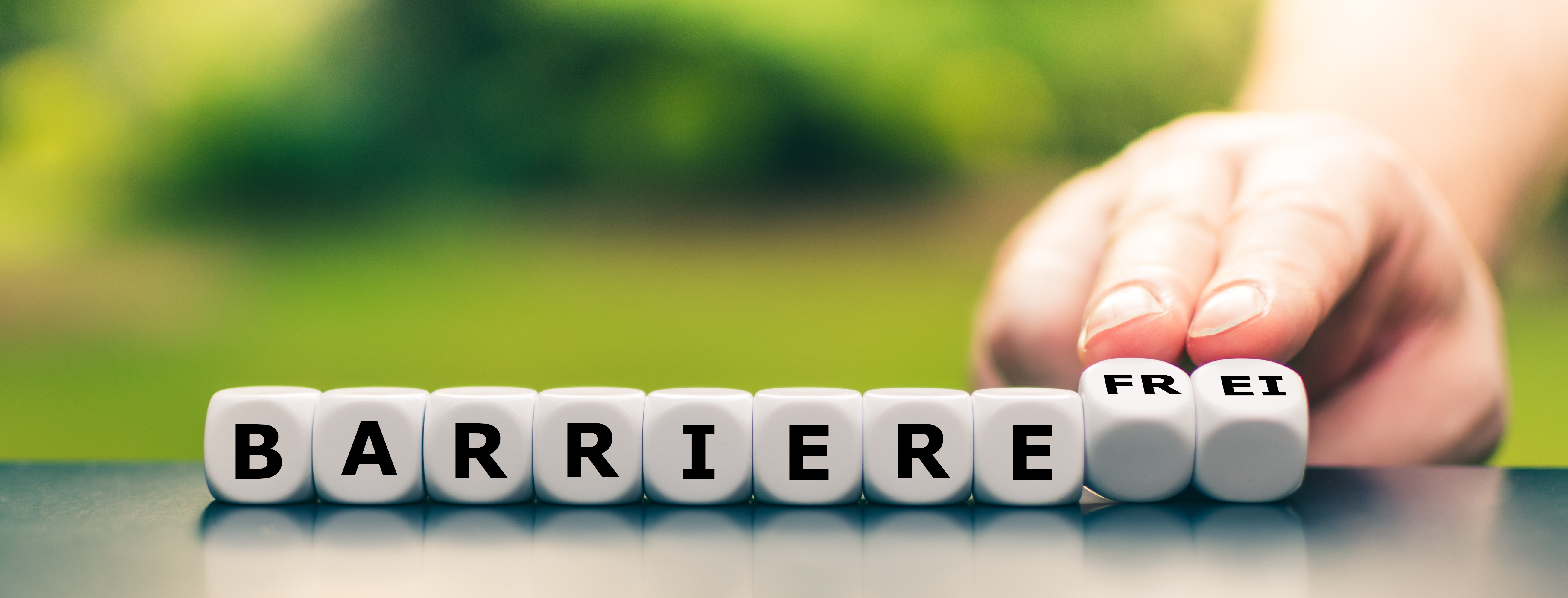 Würfel, mit Buchstaben, die nebeneinander aufgereiht sind und das Wort: "Barrierefreiheit" ergeben. Daneben eine Hand, die zwei der Würfel umdreht.